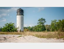 Weißer Wasserturm mit schwarzem Dach und Uhr, umgeben von Wiesen, vereinzelten Bäumen und Büschen, unter klarem, blauem Himmel.