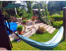 Ein Gartenbereich mit einer Steinterrasse, auf der Holzmöbel, Topfpflanzen und Hängepflanzen zu sehen sind. Im Vordergrund hängt eine blaue Hängematte, dahinter ein aufblasbarer blauer Pool und eine mit Stufen erreichbare, von Grün umgebene Sitzfläche.