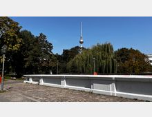 Blick auf eine weiße Brücke mit Kopfsteinpflaster und Laternen im Vordergrund, dahinter eine Baumreihe mit einer großen Trauerweide. Im Hintergrund ragt der Berliner Fernsehturm in den blauen Himmel.