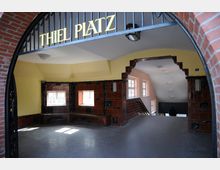 Gebäudedurchgang mit einem Schriftzug "Thiel Platz" am oberen Rand eines gemauerten Torbogens. Der Innenraum ist mit gelb gestrichenen Wänden, braunen Fliesen und Fenstern gestaltet, die natürliches Licht hereinlassen. Eine Treppe führt im hinteren Bereich nach unten.