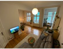 Wohnzimmer und Essbereich in einer Altbauwohnung mit Holzdielenboden, hohen Decken und großen, weiß umrahmten Fenstern. Im Vordergrund eine graue Polstercouch, ein Fernseher und Pflanzen; im Hintergrund ein Esstisch mit hängenden Lampen sowie ein Schreibtisch und Bücherregal.