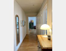 Ein schmaler Hausflur mit Holzboden und heller Wandgestaltung. Links hängt ein großer, gerahmter Spiegel sowie mehrere Bilderrahmen, rechts steht eine Kommode mit einer weißen Tischlampe. Am Ende des Flurs führt eine geöffnete Tür in einen weiteren Raum mit Regalen, Pflanzen und einem Sitzkissen.