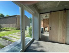 Ein Innenraum mit großen Glas-Schiebetüren, die in einen Außenbereich mit Gras und einem Gebäude im Hintergrund führen. Rechts im Bild befindet sich eine Holztür auf einer schwarzen Schiene, die zu einem Raum mit Backsteinwand und einem Waschtisch führt.