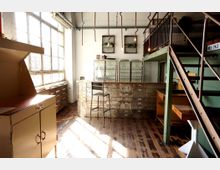 Lichtdurchfluteter Raum mit industriellem Stil, einschließlich großer Fenster, Metallregalen mit Glastüren und einer Theke aus Schubladenelementen. Im Vordergrund befindet sich ein Schrank, während eine Metalltreppe rechts zur oberen Etage führt.