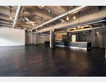 Ein offener, moderner Veranstaltungsraum mit dunklem Bodenbelag, sichtbaren Betondecken und freiliegenden Lüftungsrohren. Im Hintergrund ist ein Tresenbereich mit minimalistischer Einrichtung und indirekter Beleuchtung zu sehen.