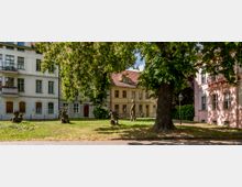 Ein kleiner Park mit einer großen Kastanie in der Mitte, umgeben von historischen Wohnhäusern in Pastellfarben. Auf der Rasenfläche stehen mehrere kleine Statuen sowie eine Laterne.