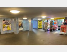 Unterführung in einem U-Bahnhof mit hellblauen Wandfliesen, Werbepostern und mehreren Leuchtkörpern an der Decke. Rechts befindet sich ein BVG-Ticketladen mit gelber Beschilderung, Verkaufsständern und einem Getränkekühlschrank.