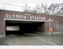 Backsteintunnel mit der Aufschrift „Zum Olympia-Stadion“, darüber ein Metallgeländer und daneben ein Wegweiser sowie ein orangefarbener Mülleimer. Im Hintergrund sind Bäume sichtbar.