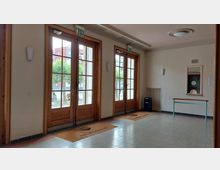 Eingangsbereich eines Gebäudes mit drei hohen Holztüren, die nach draußen führen. Innen sind Fliesenboden, eine Theaterkasse mit Glasscheibe an der rechten Wand und ein Wandlampenpaar sichtbar; vor den Türen liegen Fußmatten.