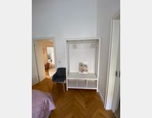 Weißer, minimalistisch gestalteter Kleiderständer mit integrierten Regalfächern in einem Raum mit Parkettboden; daneben ein grauer Polsterstuhl. Im Hintergrund sind eine offene Tür und ein angrenzender heller Raum zu sehen.