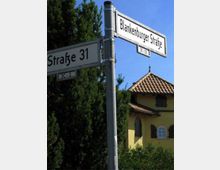 Straßenschild mit den Beschriftungen „Blankenburger Straße“ und „Straße 31“ vor einem gelben Gebäude mit roten Dachziegeln und dunkelgrünen Fensterläden, umgeben von Bäumen und Sträuchern.