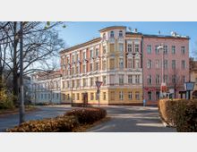 Eckhaus mit historischer Fassade, gelb und weiß gestrichen, im Stil des 19. Jahrhunderts, daneben ein rosafarbenes Gebäude. Im Vordergrund befinden sich eine Straße mit Verkehrszeichen, Fußwege und Hecken, während kahle Bäume die Szenerie säumen.