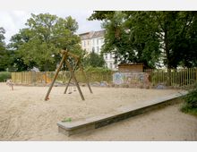 Ein Kinderspielplatz mit Sandfläche, Schaukelgestell aus Holz und weiteren Spielgeräten wie einer Rutsche im Hintergrund. Der Spielplatz ist von einer niedrigen Mauer mit Graffiti und einem Zaun umgeben; im Hintergrund sind Bäume und Wohnhäuser zu sehen.