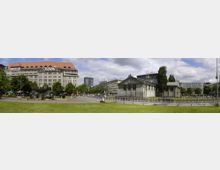 Panorama eines städtischen Platzes mit einer großen steinernen Kaufhausfassade (KaDeWe) links und einem neoklassizistischen Gebäudekomplex mit Säulen rechts, umgeben von Grünflächen, Gehwegen und vereinzelten Bäumen. Im Hintergrund sind mehrere moderne Gebäude und ein teils bewölkter Himmel zu sehen.