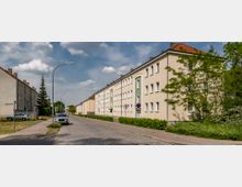 Straßenszene in einem Wohngebiet mit mehrstöckigen, hellbeigen Reihenhäusern im typisch schlichten Stil, flankiert von Gehwegen, Grünflächen und einzelnen Bäumen. Gepflasterte Fahrbahn mit vereinzelten parkenden Autos, unter blauem Himmel mit vereinzelten Wolken.