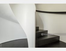 Geschwungene, weiße Wendeltreppe mit dunklen Holzstufen und schwarzem Handlauf in einem minimalistischen, hellen Innenraum. Der Boden besteht aus dunkelgrauem Material.