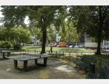 Ein kleiner Park mit Bäumen, grünen Bänken und zwei Tischtennisplatten auf einem sandigen Platz. Im Hintergrund sind eine Straße mit geparkten Autos, ein gelber Lieferwagen und Gebäude mit Geschäften zu sehen.