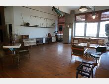 Großer Raum mit Holzboden und einer Mischung aus industriellen und rustikalen Möbeln, darunter ein langer Schrank aus recyceltem Holz und mehrere Sitzgelegenheiten. Große Fenster mit Holzjalousien lassen viel Licht herein, und die Einrichtung umfasst Details wie hängende Lampen und dekorative Gegenstände wie einen alten Globus und Vasen.