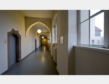 Ein langer Innenflur mit gewölbten Deckenbögen und hängenden Kugelleuchten, links eingerahmte Türen mit Steinbögen, rechts große Fenster mit Blick nach draußen auf ein weißes Gebäude mit roten Dachziegeln.