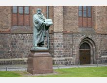 Statue von Martin Luther aus grünlicher Bronze auf einem Sockel aus rotem Stein, vor einer Fassade aus braunen und grauen Backsteinen mit Spitzbogenfenster und gotischer Tür. Im Vordergrund Rasen und gepflasterte Wege.