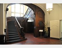 Historisches Treppenhaus mit dunkler Holzvertäfelung und geschwungener Holztreppe, die mit einem Teppichläufer bedeckt ist. Große Fenster im Hintergrund lassen Tageslicht herein, ein Kronleuchter hängt von der Decke, und rechts steht eine verzierte weiße Tür neben einer Dekoration mit getrockneten Pflanzen.