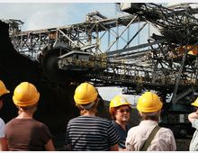 Eine Gruppe von Menschen mit gelben Schutzhelmen betrachtet einen großen industriellen Schaufelradbagger mit komplexen Stahlstrukturen und Förderbändern. Der Bagger befindet sich in einer Tagebauumgebung unter teils bewölktem Himmel.