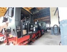 Schwarze Dampflok mit roten Akzenten in einem Lokschuppen mit Holzdach und weißen Backsteinwänden. Im Hintergrund ist ein grüner Zugwaggon zu sehen, und an der Wand hängt ein zusammengerollter Schlauch.