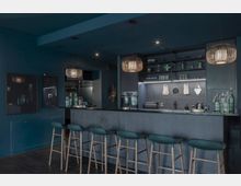 Moderne Bar in einem Innenraum mit dunkler, blauer Farbgebung. Eine Theke mit fünf hohen Hockern steht im Vordergrund, dahinter sind offene Regale, dekorative Elemente, eine Kaffeemaschine und stilvolle Hängeleuchten zu sehen.