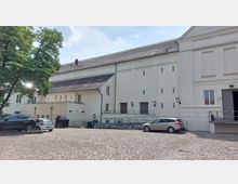 Heller Gebäudekomplex mit mehreren Etagen, weißen Fassaden und einem schrägen Ziegeldach, umgeben von einem Hof mit Kopfsteinpflaster. Im Vordergrund stehen geparkte Autos, links ein großer Baum, rechts eine Zugangstreppe und eine Rampe.