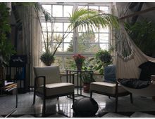 Wohnzimmer mit großen Fenstern, die einen Blick auf Bäume bieten. Der Raum ist mit zwei Sesseln, einem Tisch mit rosa Tulpen, einer Hängematte und zahlreichen grünen Zimmerpflanzen dekoriert; der Boden ist gefliest.