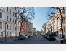 Straßenszene in einer urbanen Wohngegend mit mehrstöckigen Gebäuden, teils in hellen und teils in farbigen Fassaden. Die Straße ist von Bäumen gesäumt, mit parkenden Autos auf beiden Seiten, und eine gelbe S-Bahn fährt auf einer erhöhten Brücke im Hintergrund.