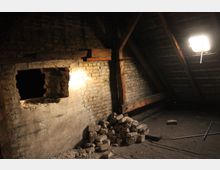 Ein verlassener Dachboden mit freiliegenden Mauersteinen und Holzbalken; ein großes Loch in der Ziegelwand und ein Haufen zerbrochener Steine auf dem Boden. Eine helle Lampe beleuchtet den Raum.