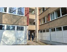 Innenhof zwischen zwei mehrstöckigen Wohnblocks mit rauer Betonfassade, weißen Fensterbändern und roten vertikalen Fassadenelementen. In der Mitte führt ein überdachter Durchgang unter dem Gebäude hindurch; unten sind große weiße Garagentore und seitlich Balkone sichtbar.