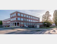 Backsteinbau mit drei Stockwerken in einem schlichten, funktionalen Baustil. Große weiße Fenster prägen die Fassade, im Vordergrund Gehwege, eine kleine Grünfläche und ein Stromkasten mit Graffiti, umgeben von Bäumen.