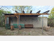 Überdachter Wartebereich im Freien mit hölzernem Dach auf Metallstützen vor einer verwitterten Betonwand mit Farbresten. Auf dem gepflasterten Boden stehen mehrere Bänke und zwei Abfallbehälter, umgeben von Bäumen und Sträuchern.