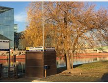 Ein Flussufer mit einem Baum, dessen gelbbraune Äste herabhängen, im Vordergrund. Im Hintergrund sind moderne Glasgebäude, eine Brücke und Treppen zu sehen, daneben ein kleines Ticket- und Informationshäuschen mit der Aufschrift „Reederei Riedel“.