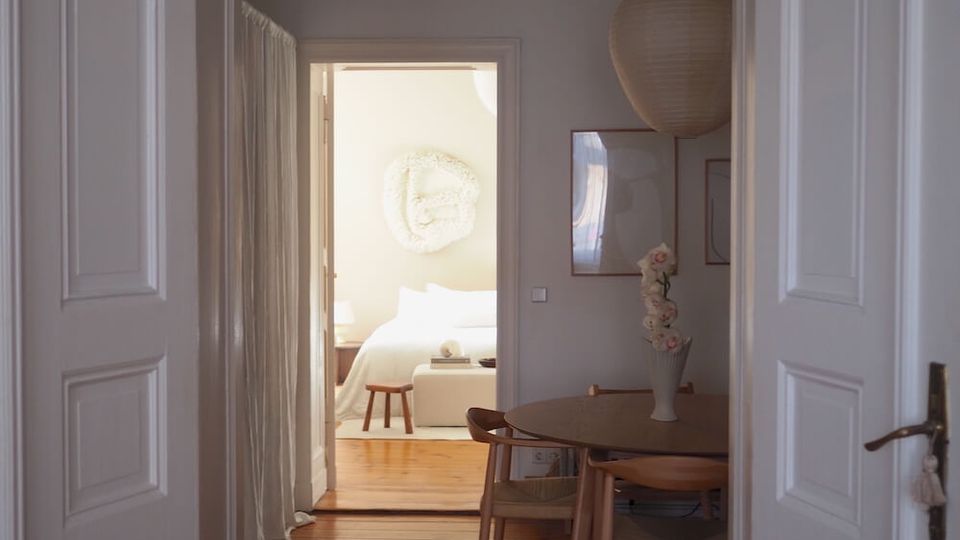View through white paneled double doors into a narrow apartment hallway with wooden floors, leading to a bright bedroom. A small dining nook with a round table, chairs, framed wall art, and a hanging paper lantern sits on the right, with a curtain along the left wall.