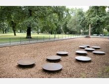 Spielplatz mit Holzschnitzelboden, runden Metallplattformen zum Balancieren und umliegendem grünen Park mit großen Bäumen im Hintergrund. Ein niedriger Zaun trennt den Spielbereich von einem Gehweg.
