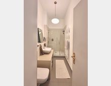 Schmales, modernes Badezimmer mit weiß gestrichenen Wänden. Im Vordergrund eine Toilette und ein Waschbecken auf einer beigen Konsole, im Hintergrund eine gläserne Duschkabine. Ein Wandheizkörper und ein heller Läufer ergänzen die Einrichtung, eine Pendelleuchte hängt zentral von der Decke.