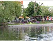 Strandbereich mit Sand und Liegestühlen am Ufer eines Gewässers, dahinter ein Holzpavillon mit Strohdach-Sonnenschirmen, umgeben von Bäumen und Gebäuden im Hintergrund.