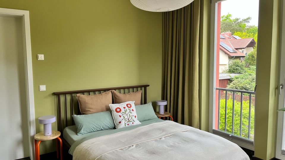 Schlafzimmer mit olivgrünen Wänden, Doppelbett mit dunkel gerahmtem Kopfteil und zwei kleinen Nachttischen. Rechts führt eine große Glastür mit Vorhang auf einen Balkon und bietet Blick auf Bäume und benachbarte Häuserdächer.