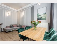 Heller, modern eingerichteter Wohn- und Essbereich mit grauen Wänden samt Stuckleisten und Wandpaneelen, einem Ecksofa und einem Holzesstisch mit grünen Polsterstühlen. An der Wand hängen ein Landschaftsbild und darunter ein Heizkörper, daneben ein großes Fenster mit dunklen Vorhängen.