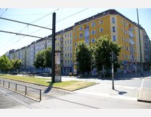Straßenecke in einer Stadt mit mehrstöckigen Wohnhäusern in gelben und weißen Fassaden. Im Vordergrund Straßenbahngleise, Grünfläche, Bäume und ein Fußgängerweg sowie Werbesäulen und Ampeln an der Kreuzung.