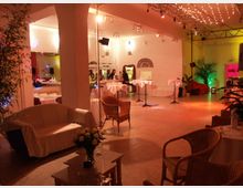 Saal mit gemütlicher Lounge-Atmosphäre, ausgestattet mit weißen Sofas, geflochtenen Stühlen und dekorativen Pflanzen. Eine Bar sowie Stehtische mit weißen Decken und dezente Beleuchtung mit warmen Farben und Lichterketten schaffen eine einladende Umgebung.