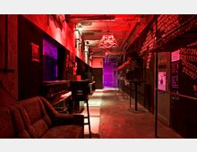 Ein schmaler, industriell gestalteter Innenraum mit roter und violetter Beleuchtung, rohen Betonwänden und dekorativen Elementen wie Wandgrafiken mit Binärcode-Mustern. Der Raum enthält eine Sitzcouch, Hocker, Hängestangen und eine Tür mit der Aufschrift „Garderobe“.