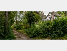 Ein schmaler, unbefestigter Weg durch eine bewaldete Umgebung mit dichter Vegetation; im Hintergrund ist ein weißes Haus mit Satteldach und Balkon zu sehen, umgeben von Bäumen.