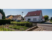 Wohnsiedlung mit Einfamilienhäusern, die weiße und gelbe Fassaden sowie rote und dunkle Dachziegel haben. Im Vordergrund ein kleiner Garten mit Gemüsebeeten, umgeben von einem Metallzaun, und eine gepflasterte Straße mit Gehwegen.