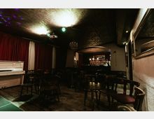 Dimly lit lounge or small music venue interior with round tables and wooden chairs on a dark wood floor, a white upright piano by red curtains, and a bar with bottles under an arched opening. Low ceiling lighting and a chandelier add to the intimate, basement-like setting.