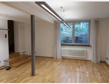 Ein leerer Raum mit hellem Parkettboden, großen Fenstern mit weißen Vorhängen und einem Blick auf eine Steinmauer mit bepflanzten Blumentöpfen. In der Ecke befinden sich Holzstufen, die zu einem angrenzenden Bereich führen, sowie ein Heizkörper unter den Fenstern.