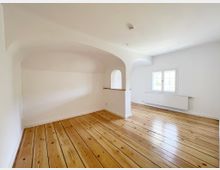 Heller Raum mit weißen Wänden und hellem Holzfußboden, einem kleinen Rundbogen in der Wand und einem großen Sprossenfenster, das Tageslicht hereinlässt. Ein Heizkörper befindet sich unter dem Fenster, und die Decke ist schlicht gestaltet.